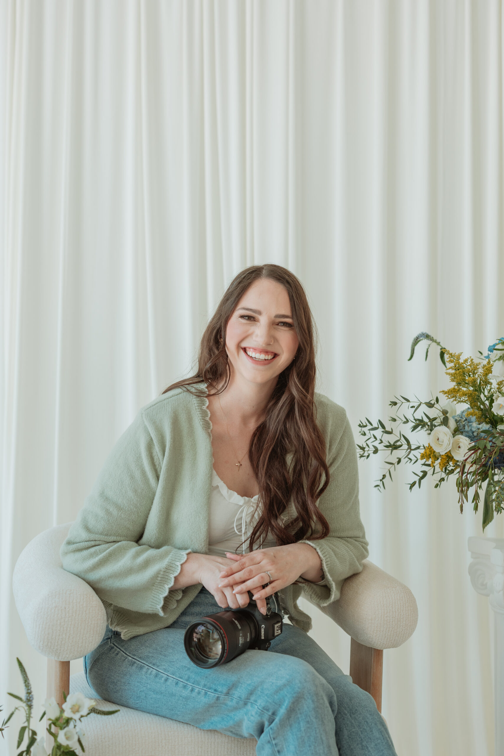 Kayla, owner and photographer behind Wren & Elm Photography, smiling in a bright studio beside floral arrangements